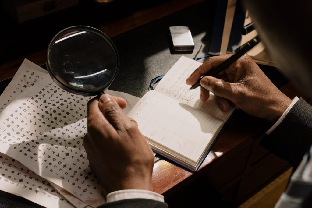A man in a suit examines a book and pen closely with a magnifying glass.