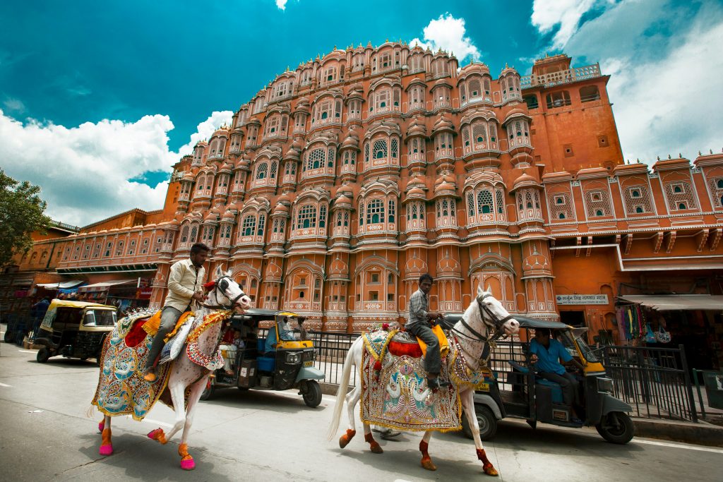 a man riding horses in front of a large building with Hawa Mahal in the background
