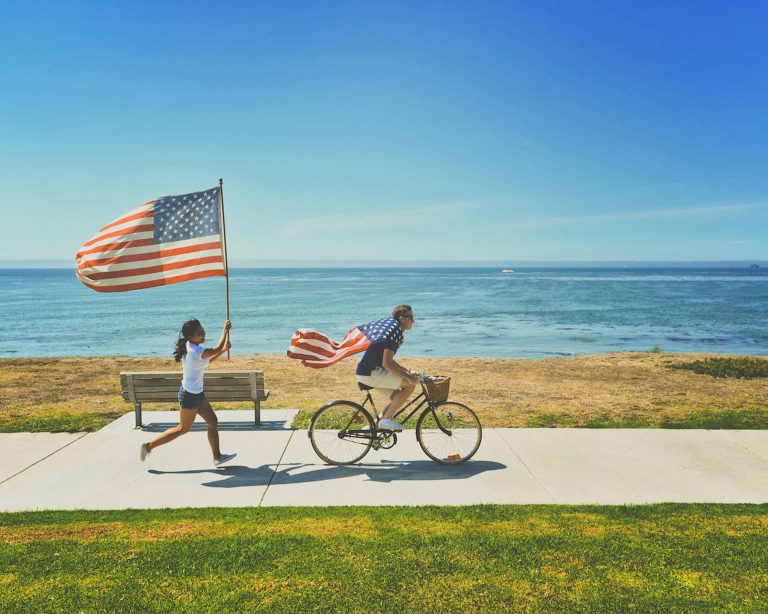 a man and woman riding a bicycle with a flag and a woman running on a bench