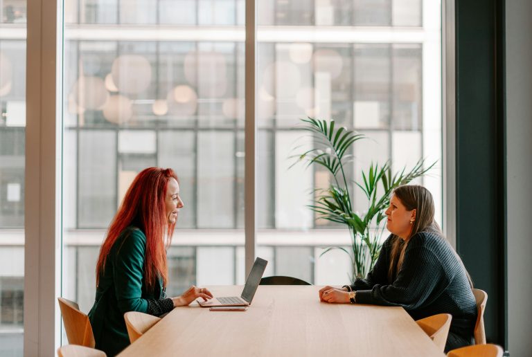 two women sitting at a table looking at a laptop