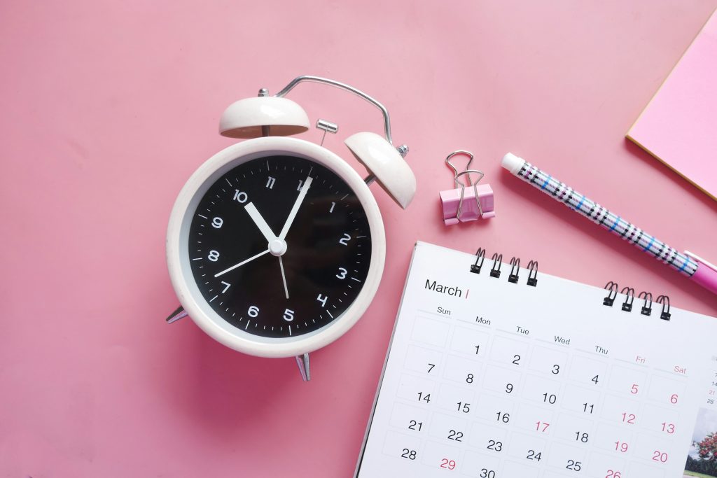 a white alarm clock next to a calendar and a pen